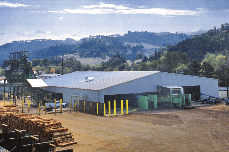 Metal industrial building at a lumber processing site, with stacks of logs in the foreground, open equipment bays, and wooded hills rising in the background.